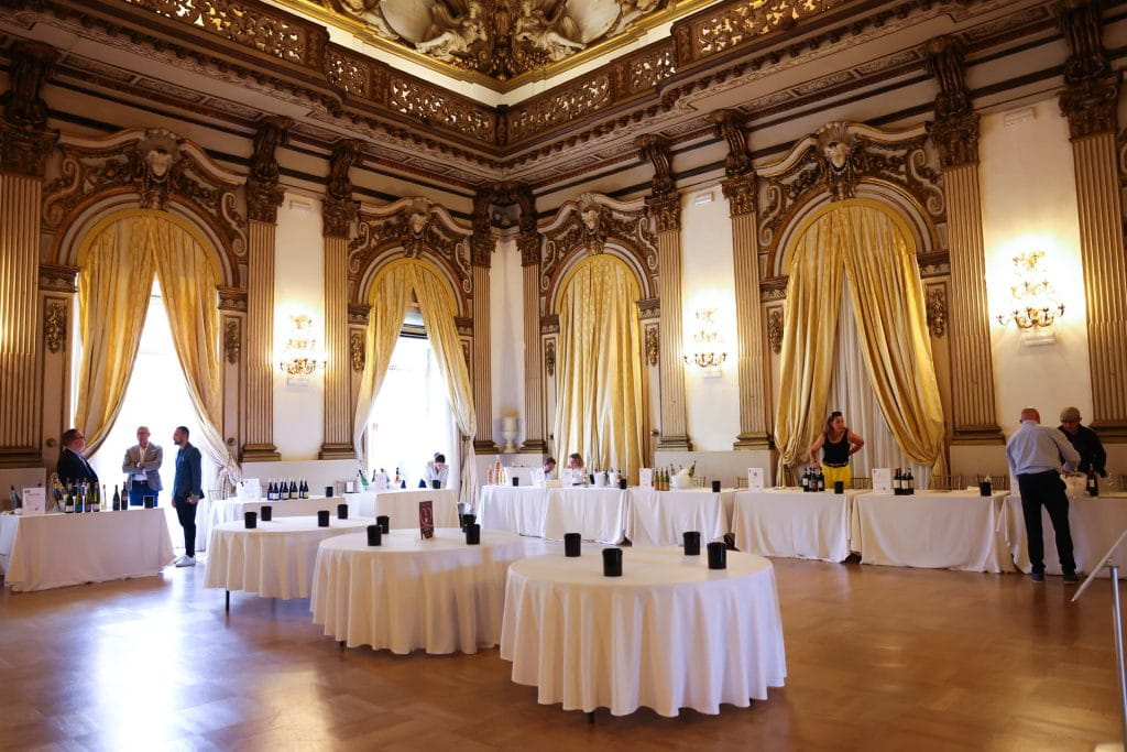 Elegante sala da ballo con dettagli dorati, soffitti alti e grandi finestre con tende. Diverse persone sono in piedi vicino a tavoli coperti da teli bianchi che espongono bottiglie, probabilmente per una degustazione o un'esposizione.
