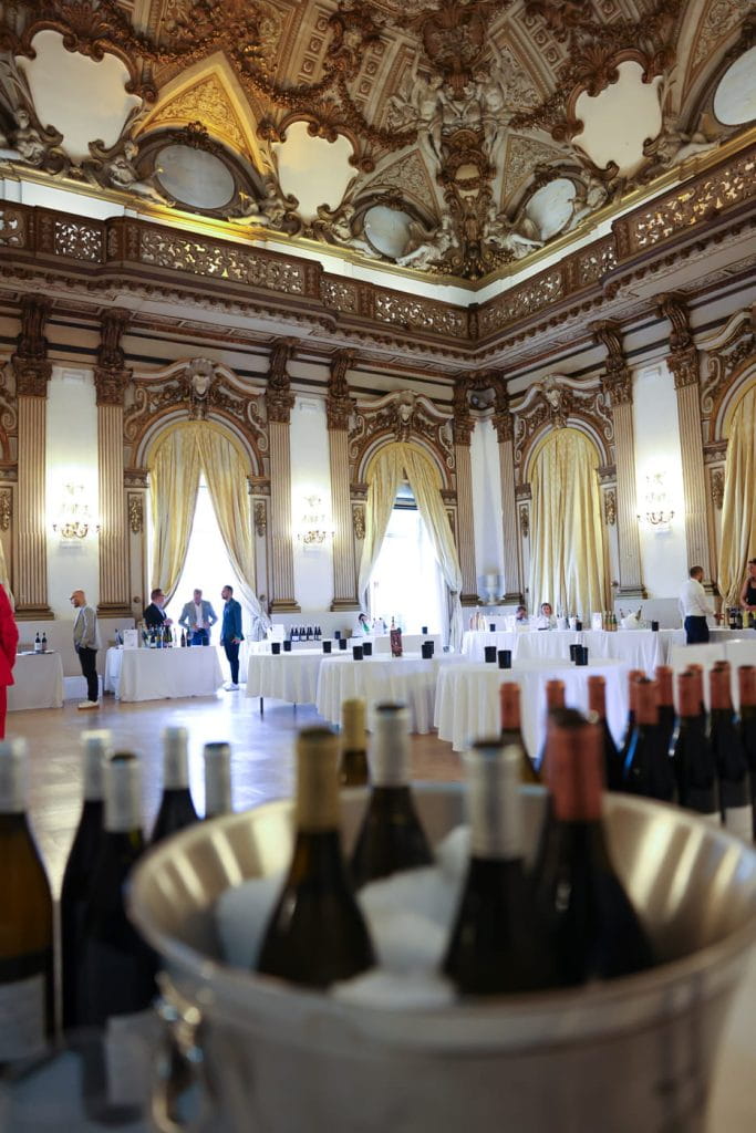 Una grande sala decorata con soffitti alti e dettagli dorati ospita una degustazione di vini. Tavoli con bottiglie sono allineati nella stanza e le persone conversano in piedi. In primo piano, le bottiglie di vino sono raffreddate in un grande secchio di ghiaccio.
