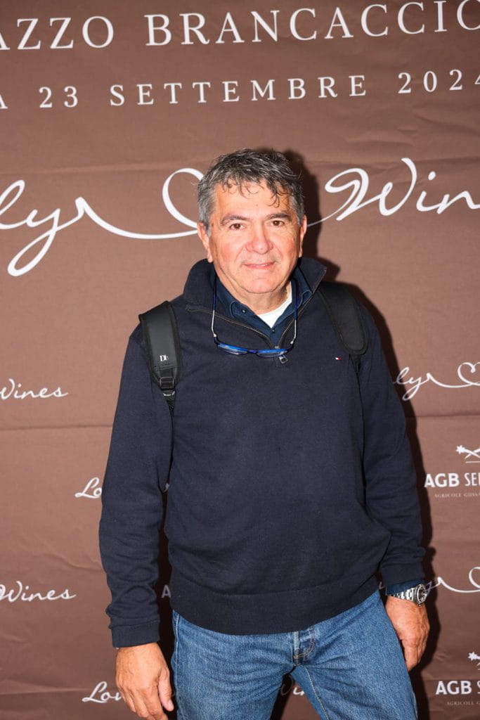 Un uomo con i capelli corti e grigi, che indossa un maglione blu e jeans, è in piedi davanti a un fondale marrone con testo bianco che promuove un evento a Palazzo Brancaccio il 23 settembre 2023. Ha uno zaino e sorride.