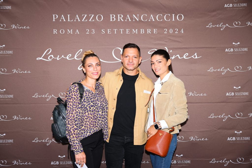 Tre persone sono in piedi e sorridono durante un evento fotografico davanti a un fondale brandizzato con la scritta Palazzo Brancaccio, Roma 23 settembre 2024 e con testi e loghi decorativi relativi a Lovely Wines.