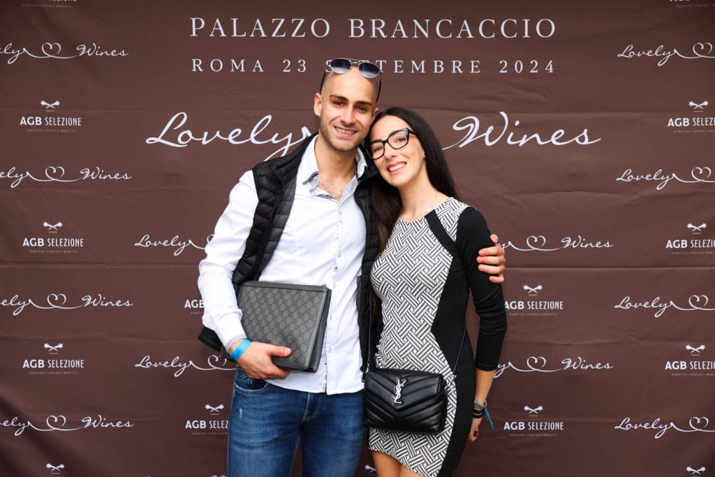 Un uomo e una donna sorridenti stanno a braccetto davanti a un fondale per un evento Lovely Wines a Palazzo Brancaccio, Roma, 23 settembre 2024. L'uomo tiene in mano una cartella trapuntata; entrambi sono vestiti in modo elegante.