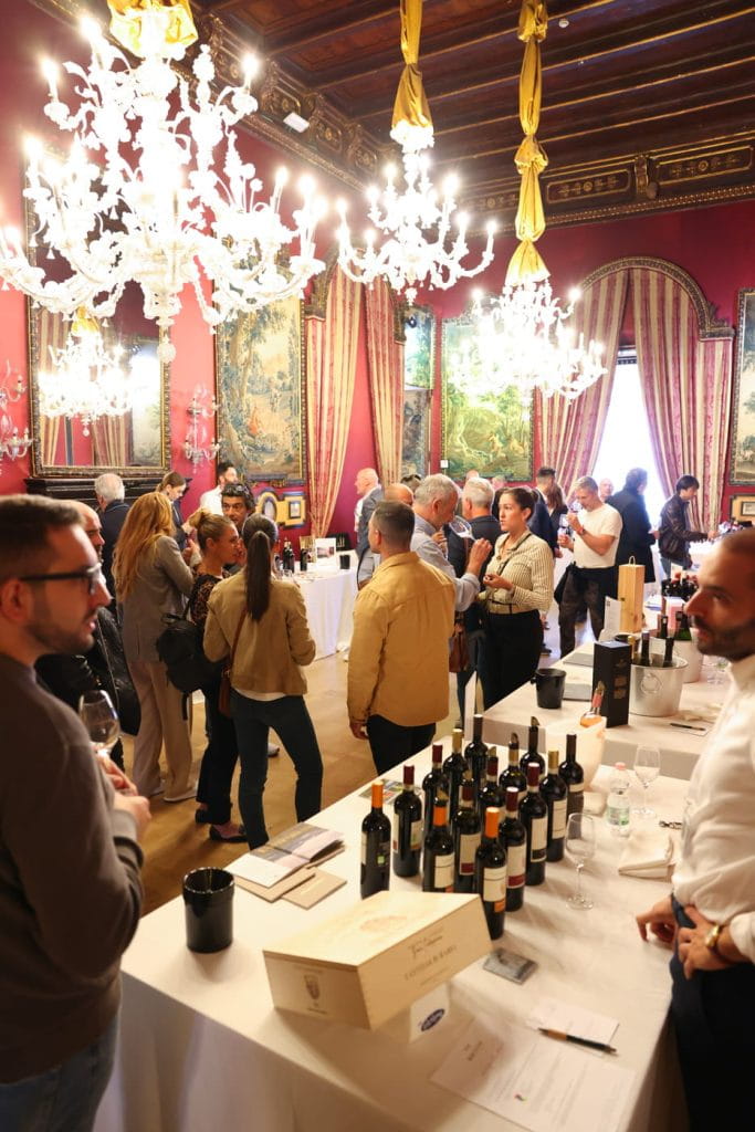 Un gruppo di persone si riunisce in una sala ornata e illuminata da lampadari con pareti rosse e alte finestre, degustando vino su diversi tavoli coperti di bottiglie e note di degustazione.