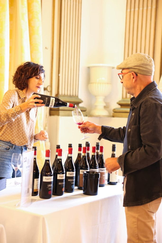 Una donna versa del vino rosso in un bicchiere tenuto in mano da un uomo durante una degustazione di vini. Sul tavolo tra i due sono esposte diverse bottiglie di vino. L'ambiente sembra essere al chiuso con un arredamento classico.