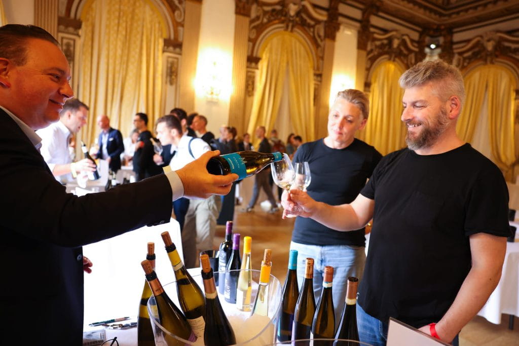 Un uomo versa del vino bianco nel bicchiere di un altro uomo durante una degustazione di vini in una sala elegante con decorazioni e tende dorate. Sullo sfondo, diverse persone assaggiano i vini e conversano.