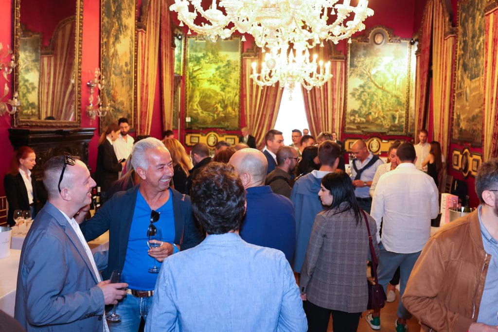 Un gruppo di persone sociali in un evento al chiuso, in una sala ornata con pareti rosse, lampadari e grandi arazzi incorniciati. Alcuni tengono in mano dei drink mentre si mescolano e conversano.
