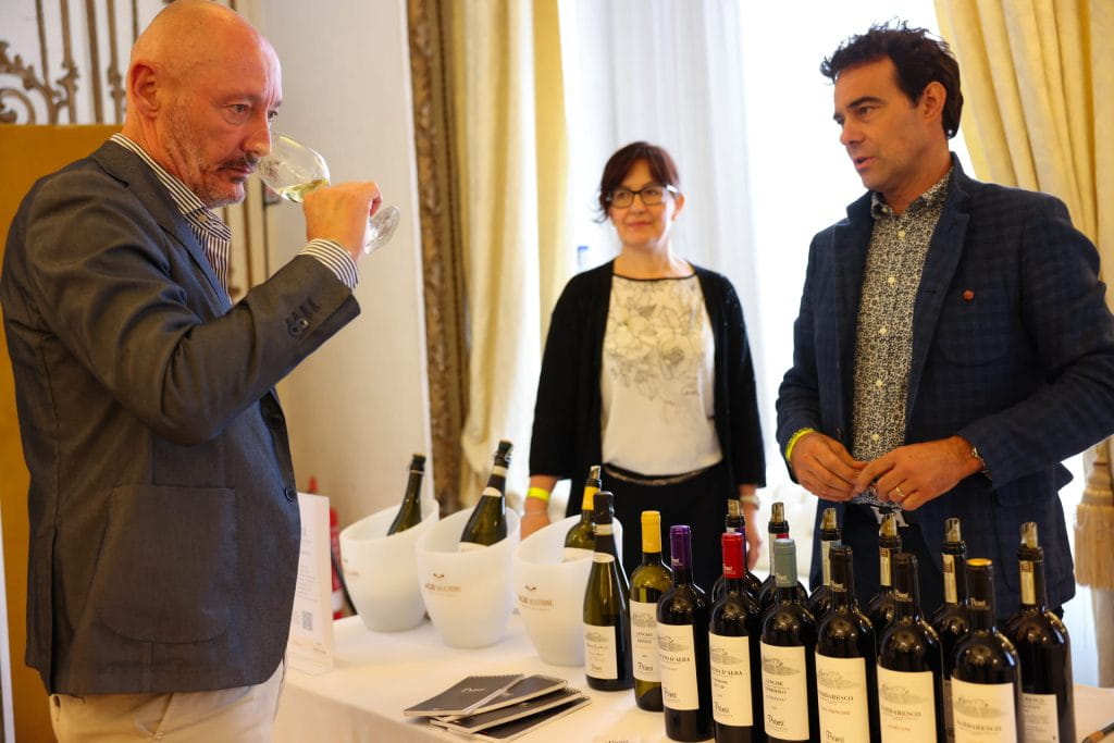 Un uomo in giacca e cravatta degusta del vino su un tavolo che espone diverse bottiglie di vino, mentre un altro uomo e una donna stanno dietro il tavolo, probabilmente per presentare i vini. Sullo sfondo sono visibili tende bianche e secchielli per il ghiaccio.