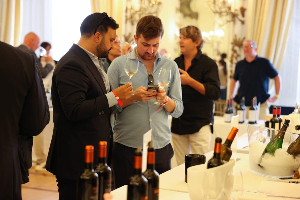 Due uomini in piedi a un evento di degustazione di vini, con in mano bicchieri e bottiglie. Uno controlla il suo telefono mentre altri, sullo sfondo, conversano. L'ambiente è elegante, con luci intense e bottiglie di vino esposte sui tavoli.