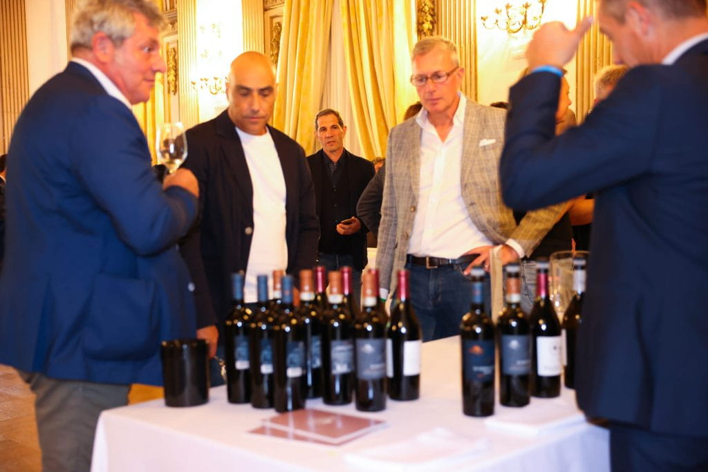 Un gruppo di uomini si trova intorno a un tavolo con diverse bottiglie di vino in occasione di un evento al chiuso, impegnato in una conversazione e in una degustazione di vini in una stanza elegante e ben illuminata.