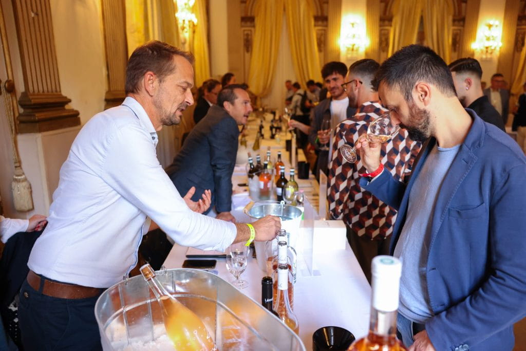 Le persone partecipano a una degustazione di vini in una sala elegante con soffitti alti e tende dorate; un uomo dietro il tavolo versa il vino mentre un altro annusa un bicchiere. Bottiglie e bicchieri sono disposti sul tavolo.