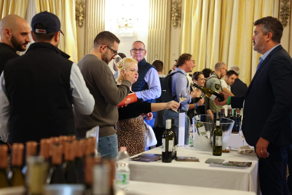Le persone partecipano a un evento di degustazione di vini all'interno, assaggiando e versando il vino su tavoli allineati con le bottiglie. La sala è caratterizzata da eleganti tende gialle e da pareti bianche decorate.
