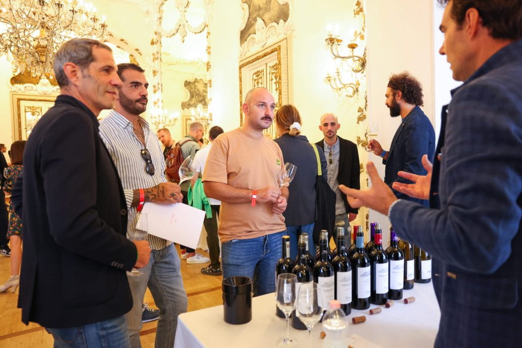 Un gruppo di persone si trova intorno a un tavolo con bottiglie e bicchieri di vino durante una degustazione al chiuso. Un uomo sta spiegando qualcosa mentre gli altri ascoltano con attenzione, tenendo in mano dei bicchieri di vino. La stanza è elegante, con lampadari e pareti decorate.