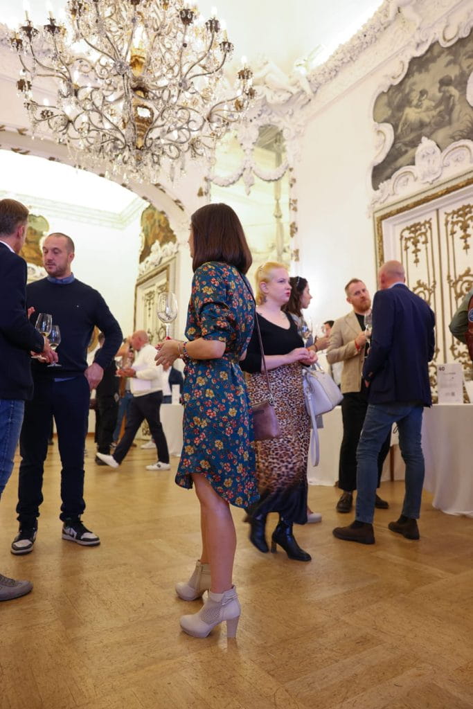 Un gruppo di persone socializza in un elegante evento al chiuso con pareti bianche decorate, decorazioni dorate e un grande lampadario. Diversi ospiti bevono e conversano in piccoli gruppi su un pavimento di legno lucido.