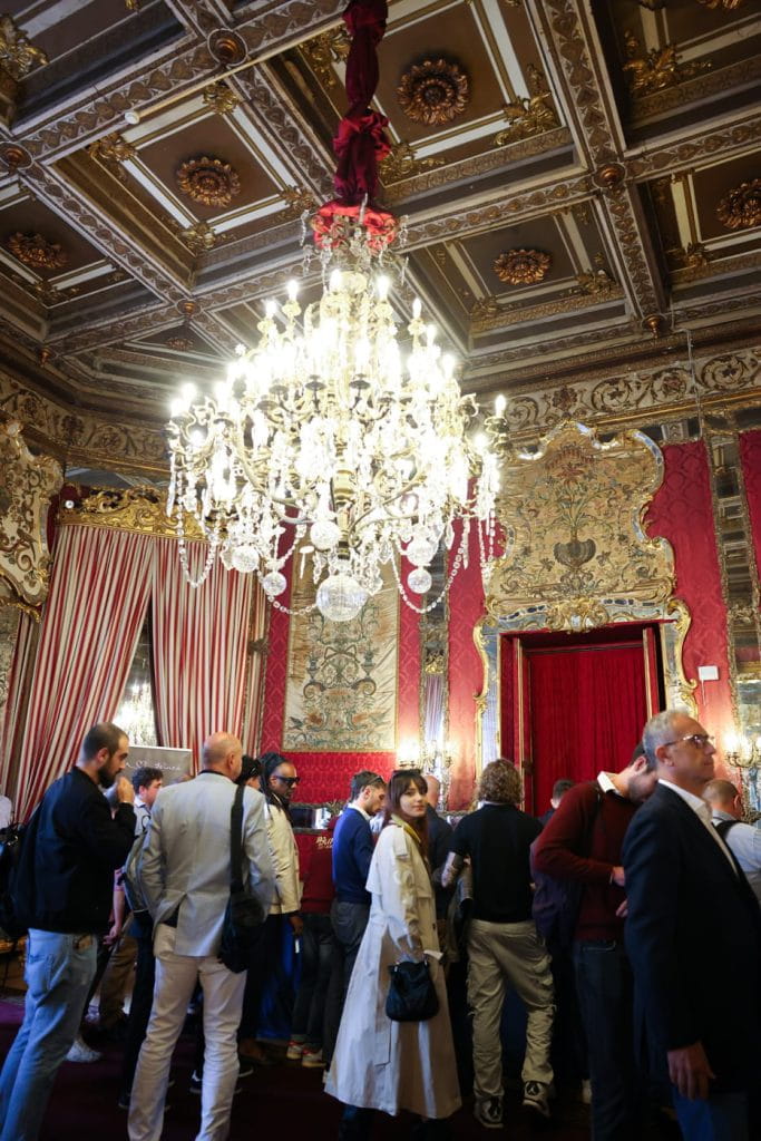 Un gruppo di persone si trova in una stanza decorata con pareti rosse, decorazioni dorate e un grande lampadario di cristallo che pende da un soffitto intricato. Tende a righe e arredi lussuosi contribuiscono a creare un'atmosfera elegante.