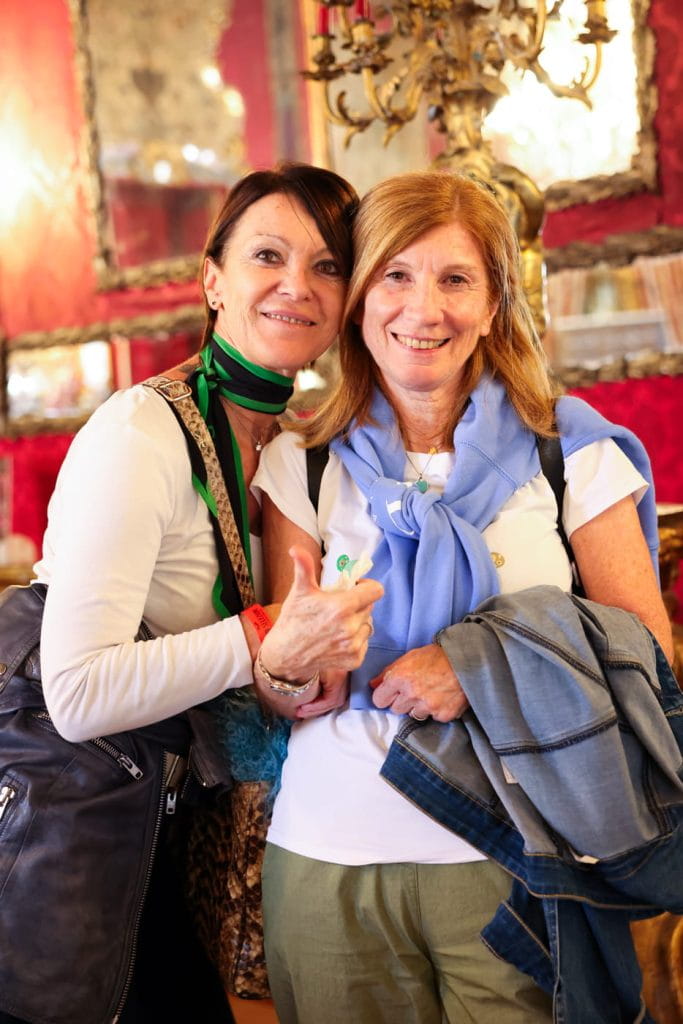 Due donne sorridono e posano insieme in una stanza caldamente illuminata e decorata con pareti rosse, specchi e una lampada decorativa sullo sfondo. Una delle due donne alza il pollice.