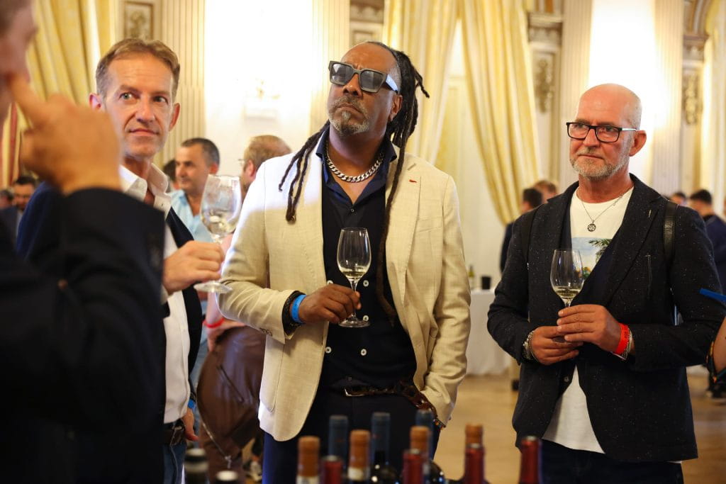 Tre uomini si trovano insieme a un evento di degustazione di vini, ciascuno con un bicchiere di vino in mano. Sono vestiti in modo elegante e casual, con bottiglie di vino in primo piano e un arredamento elegante sullo sfondo.