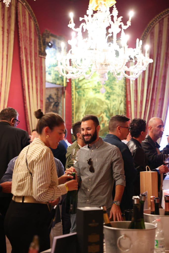 Un gruppo di persone socializza in un elegante evento al chiuso con lampadari e drappi decorati. Un uomo e una donna sorridenti parlano vicino a un tavolo con bottiglie e secchielli per il ghiaccio. L'atmosfera è vivace e festosa.