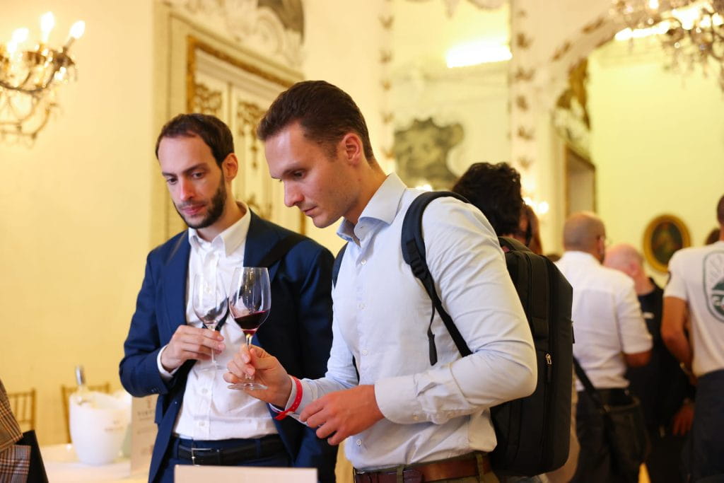 Due uomini in abiti formali si trovano al chiuso durante un evento elegante, ognuno dei quali tiene in mano un bicchiere di vino rosso e lo esamina attentamente. Sullo sfondo sono visibili lampadari, decorazioni e altre persone.