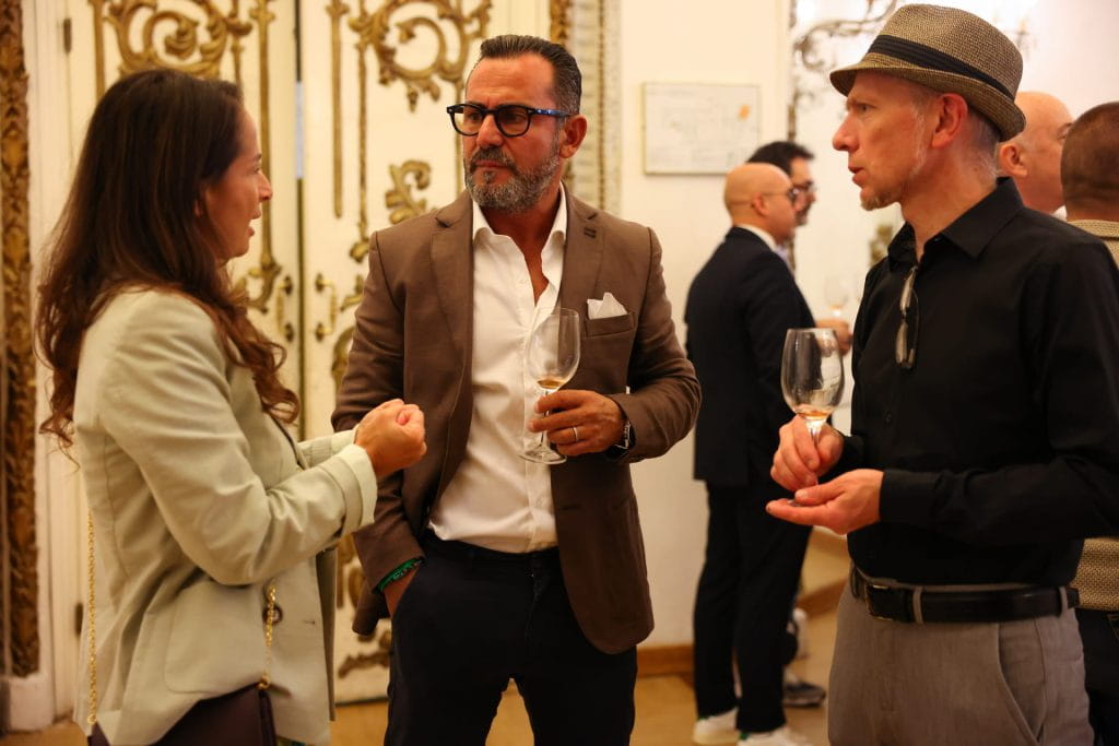 Tre persone in abiti formali parlano durante un evento sociale al chiuso, tenendo in mano un bicchiere di vino. Sullo sfondo sono presenti porte ornate in bianco e oro e altri ospiti sono visibili nella stanza.
