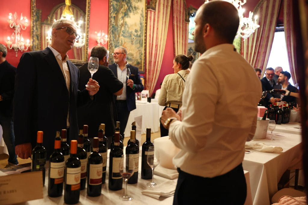 Persone a un evento di degustazione di vini in una sala elegante e decorata. Bottiglie di vino sono esposte sui tavoli e due uomini in primo piano conversano, uno dei quali tiene un bicchiere di vino. Altri ospiti si mescolano sullo sfondo.