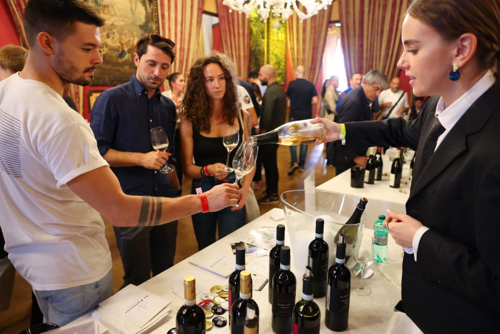 Una donna in giacca e cravatta versa del vino nel bicchiere di un uomo durante una degustazione, mentre altre tre persone guardano e tengono in mano dei bicchieri da vino. Sul tavolo ci sono diverse bottiglie di vino e un secchiello per il ghiaccio. La stanza è elegante, con lampadari e tende.