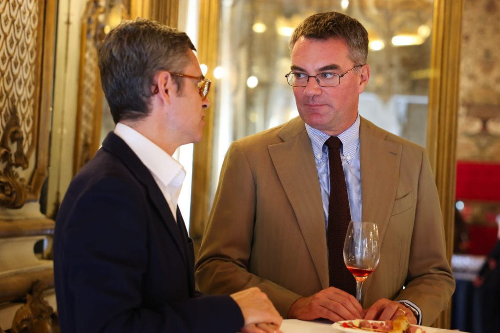 Due uomini in giacca e cravatta stanno a un tavolo e conversano. Uno tiene in mano un bicchiere di vino e un piatto con del cibo. Sullo sfondo sono visibili decorazioni dorate e uno specchio, che suggeriscono un ambiente interno elegante.