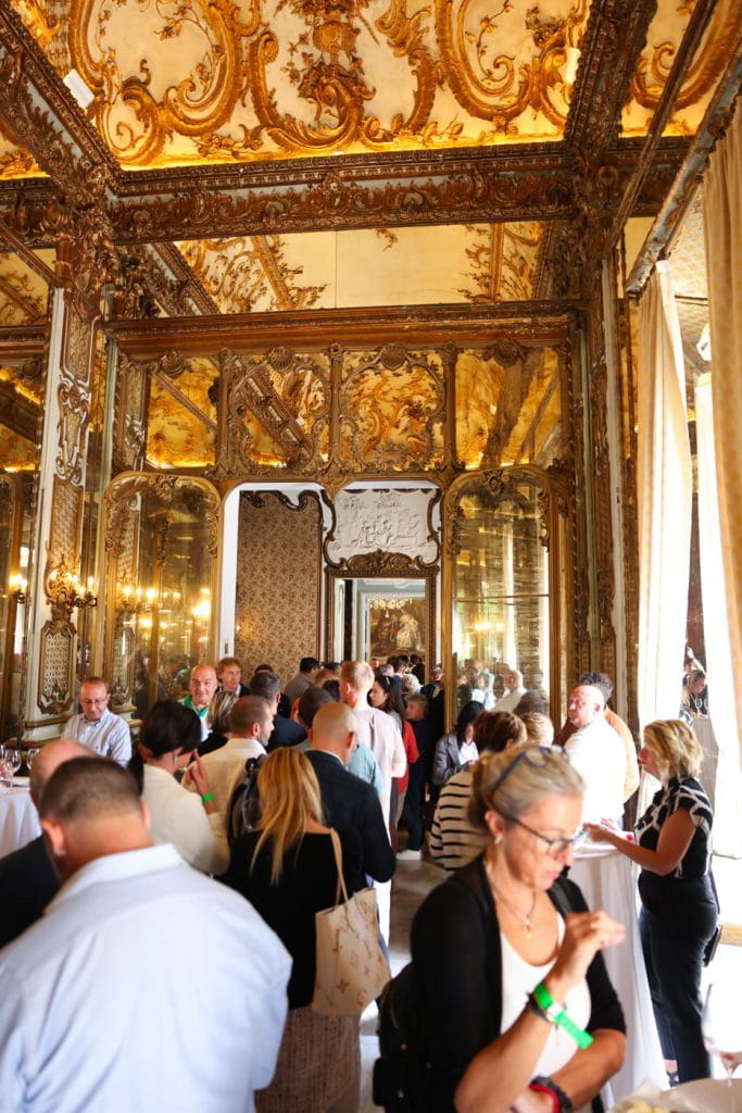 Un gruppo di persone si riunisce in una sala ornata e dorata, con elaborate decorazioni in oro, specchi e soffitti alti, alcune mescolandosi e altre mangiando o bevendo. La luce naturale entra da alte finestre con tende aperte.