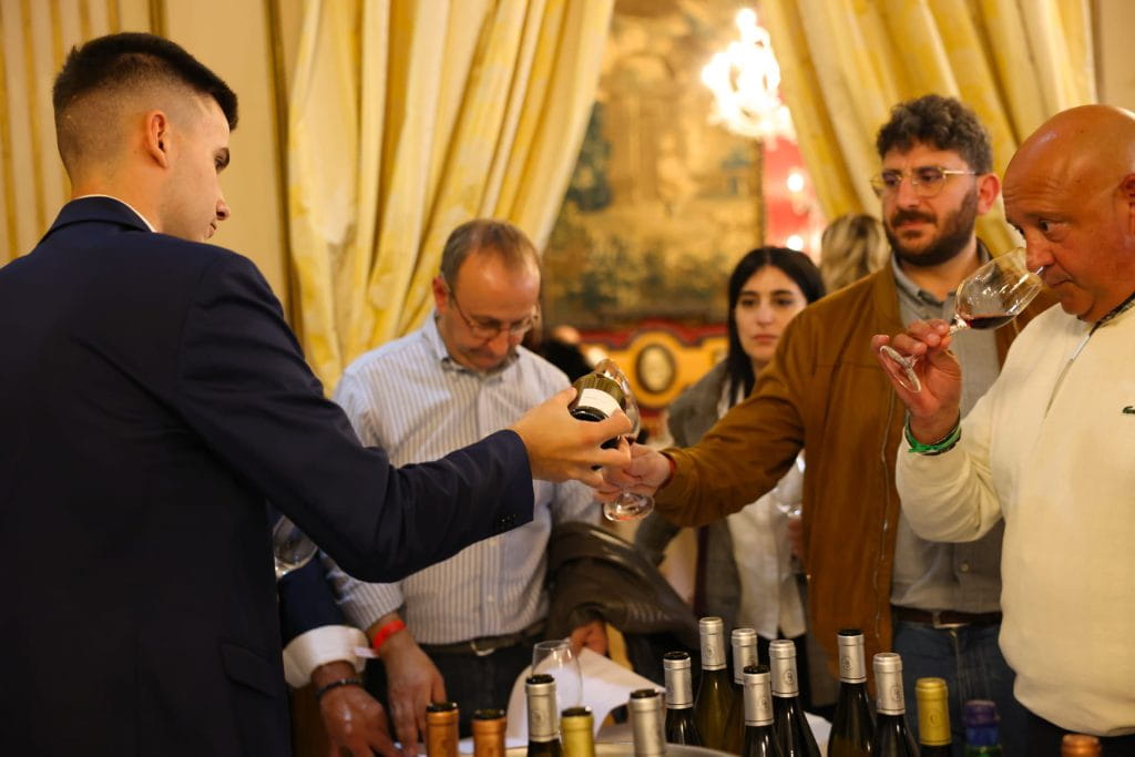 Un uomo in giacca e cravatta versa del vino in un bicchiere per un altro uomo durante una degustazione di vini, con diverse persone che osservano e bottiglie disposte su un tavolo di fronte a loro. L'ambiente appare elegante con tende drappeggiate.