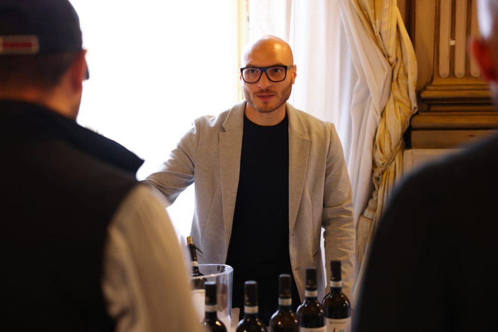 Un uomo con gli occhiali e un blazer grigio chiaro sta dietro un tavolo con diverse bottiglie di vino e parla con due persone in primo piano durante un evento al chiuso. La luce entra da una finestra alle sue spalle.