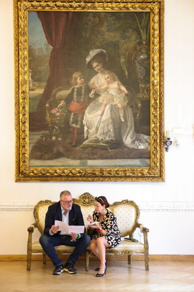 Un uomo e una donna siedono su un divano d'epoca ornato e leggono insieme dei documenti in una stanza elegante. Sopra di loro è appeso un grande quadro con cornice dorata che raffigura una donna e due bambini in abiti d'epoca.