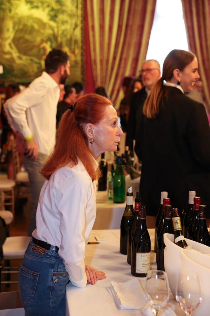 Una donna con i capelli rossi, in camicia bianca e jeans, si appoggia a un tavolo con diverse bottiglie di vino in un affollato evento al chiuso, con persone che socializzano sullo sfondo.