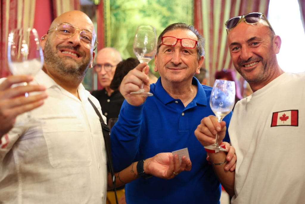 Tre uomini sorridenti, ciascuno con un bicchiere di vino in mano, sono vicini in un interno. L'uomo al centro indossa una camicia blu. Sullo sfondo sono visibili tende e persone a strisce rosse e crema.