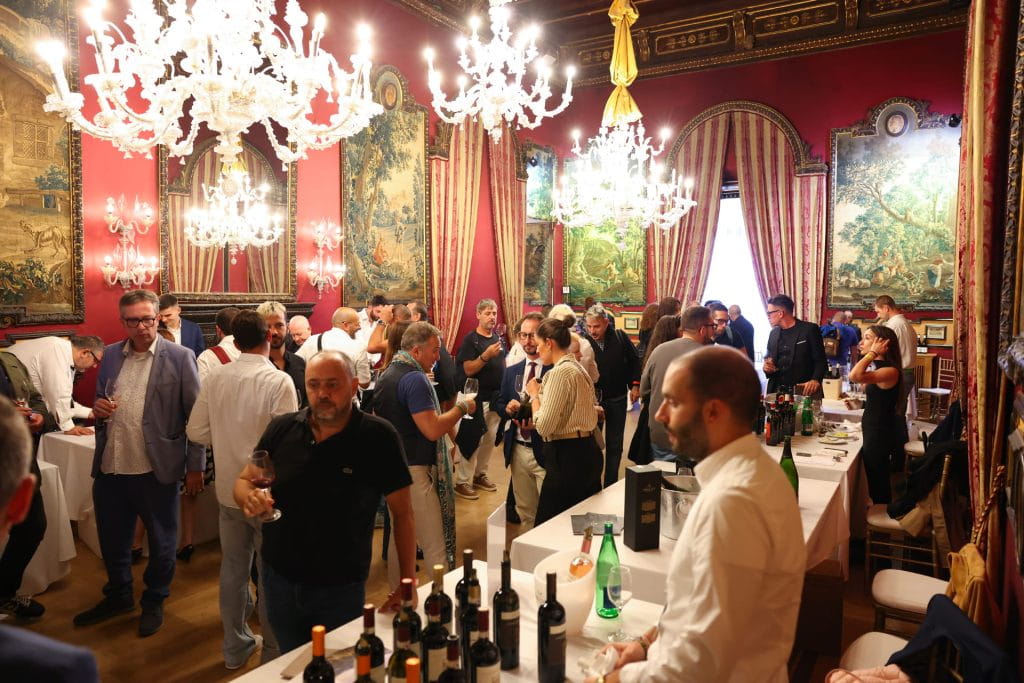 Un gruppo di persone socializza e degusta vino in una sala ornata, illuminata da lampadari, con pareti rosse, grandi quadri e alte finestre drappeggiate da tende. Sui tavoli sono disposte bottiglie e bicchieri di vino.