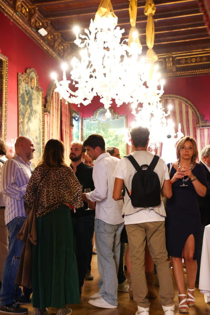 Un gruppo di persone si riunisce e conversa in una sala elegante con pareti rosse decorate, grandi lampadari e decorazioni dorate. Alcuni hanno in mano dei drink e l'atmosfera appare vivace e sociale.