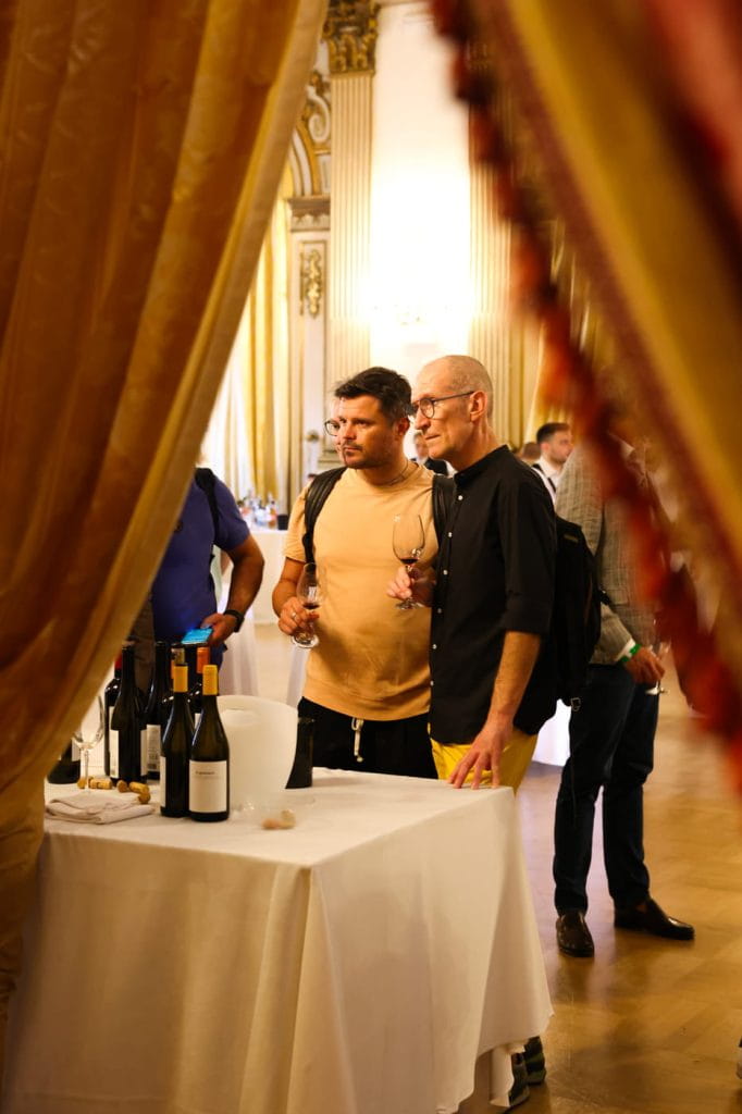 Due uomini si trovano accanto a un tavolo con bottiglie e bicchieri di vino durante un elegante evento al chiuso. Uno tiene un bicchiere di vino e l'altro gli sta vicino. Sullo sfondo sono visibili tende ornate e decorazioni dorate.