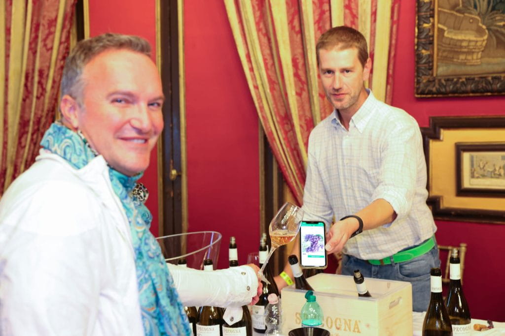 Due uomini sono in piedi accanto a un tavolo con diverse bottiglie di vino. Un uomo sorride alla telecamera mentre l'altro versa il vino e mostra un telefono con un'immagine. L'ambiente è caratterizzato da pareti rosse e tende decorative.