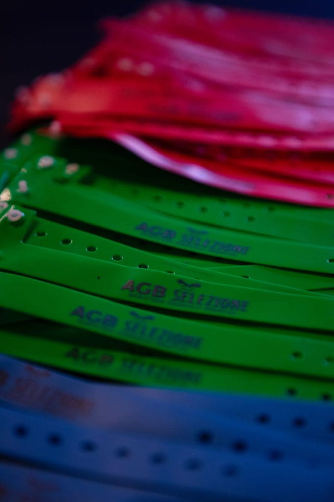 Vista ravvicinata di braccialetti di plastica impilati nei colori verde, blu e rosa, con il testo AGB SELEZIONE stampato su di essi. I braccialetti sono disposti in file sovrapposte.
