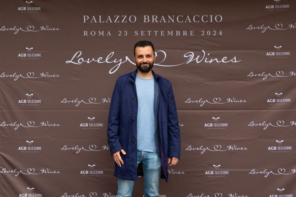 Un uomo con la barba, in cappotto blu e jeans, si trova davanti a un fondale a gradini e ripetizioni per un evento chiamato Lovely Wines a Palazzo Brancaccio, Roma, il 23 settembre 2024.