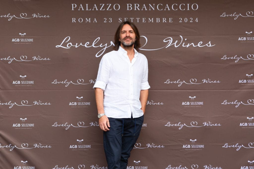 Un uomo con capelli castani lunghi fino alle spalle e barba, che indossa una camicia bianca e pantaloni scuri, è in piedi davanti a un fondale marrone con Lovely Wines e dettagli dell'evento, tra cui Palazzo Brancaccio, Roma 23 Settembre 2024.