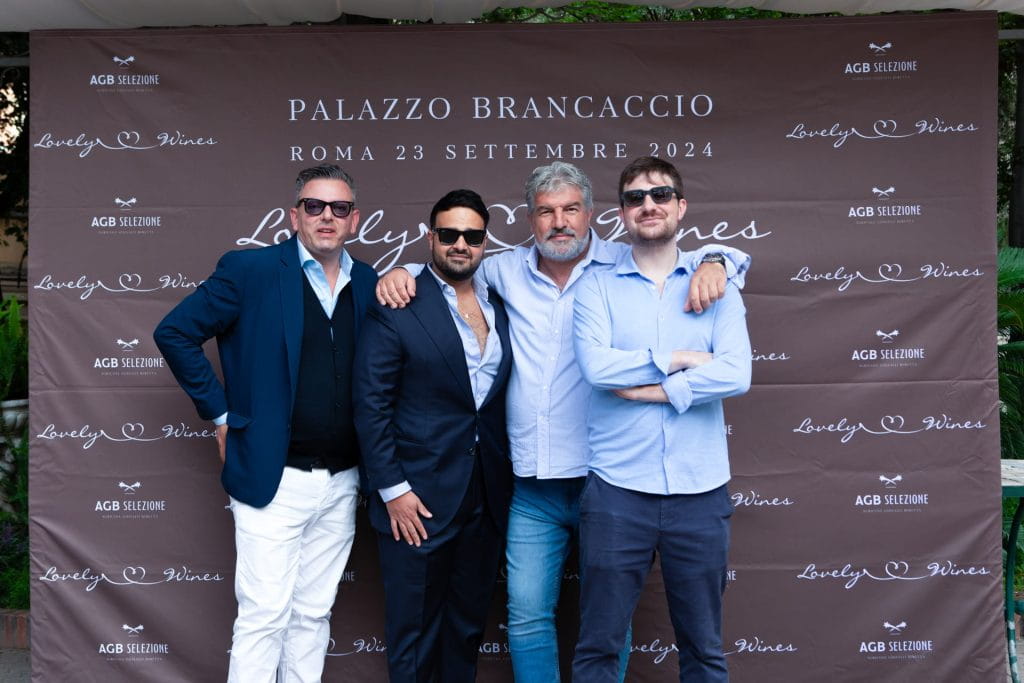 Quattro uomini con gli occhiali da sole posano insieme davanti a uno sfondo di un evento con marchio che recita "Palazzo Brancaccio, Roma 23 settembre 2024" e "Lovely Wines". Sono vestiti in modo semi-formale o casual.