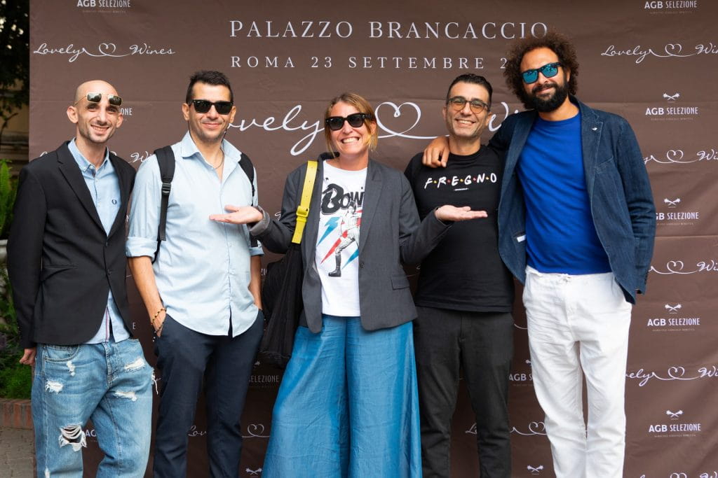 Cinque persone sorridono davanti a un fondale marrone con la scritta Palazzo Brancaccio, Roma 23 Settembre, con tre uomini che gesticolano verso una donna al centro. Sono vestiti in modo casual e indossano occhiali da sole.