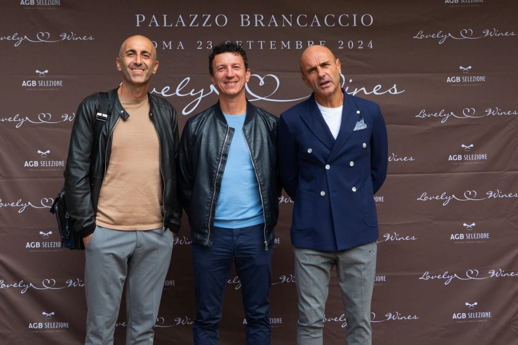 Tre uomini sono in piedi, sorridenti, davanti a un fondale marrone con testo bianco che recita "Palazzo Brancaccio, Roma, 23 settembre 2024" e "Lovely Wines". Sono vestiti con abiti casual e smart-casual.