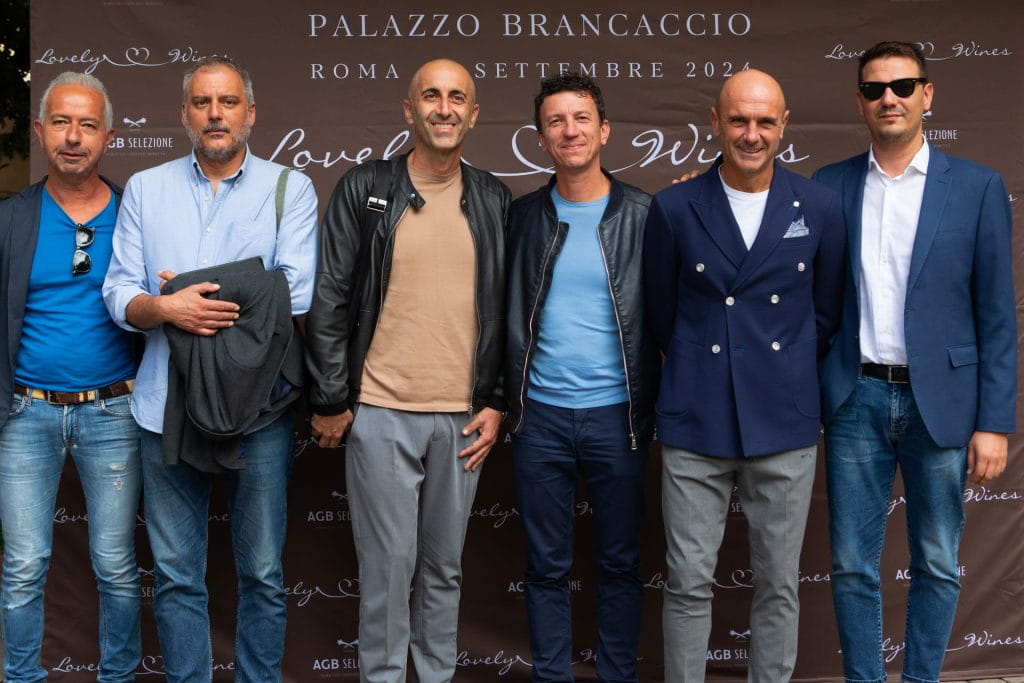 Sei uomini sono insieme e sorridenti davanti a uno sfondo con la scritta "Palazzo Brancaccio Roma Settembre 2022" e "Lovely Wines". Sono vestiti con abiti eleganti e casual, tra cui blazer e jeans.