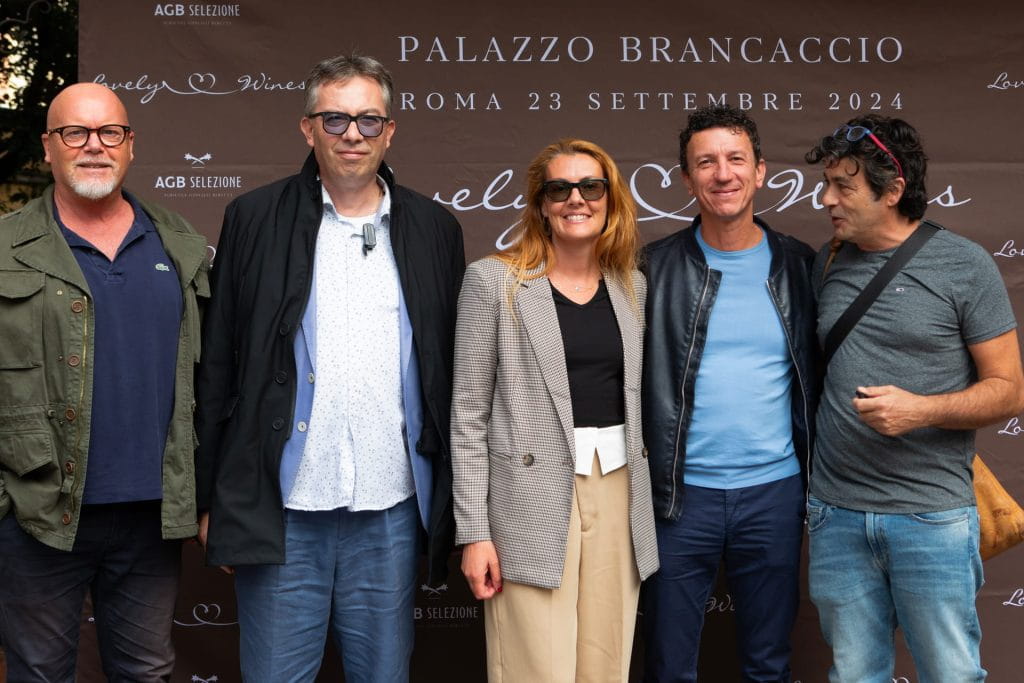 Cinque adulti sorridono insieme davanti a un fondale marrone con un testo su un evento a Palazzo Brancaccio a Roma il 23 settembre 2024. Indossano abiti casual e semi-formali.