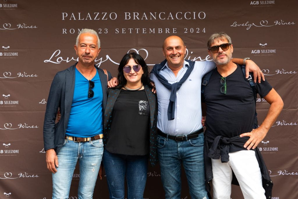 Quattro persone sono in piedi, sorridenti, davanti a uno striscione marrone con la scritta PALAZZO BRANCACCIO ROMA 23 SETTEMBRE 2024 e Lovely Wines. Sembrano partecipare a un evento e posano per una foto di gruppo.
