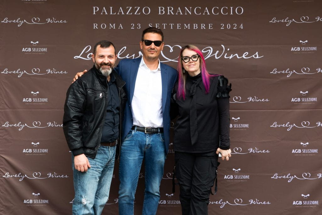 Tre persone sorridono insieme davanti a un fondale marrone con testo bianco che promuove l'evento "Lovely Wines" a Palazzo Brancaccio, Roma, il 23 settembre 2024.