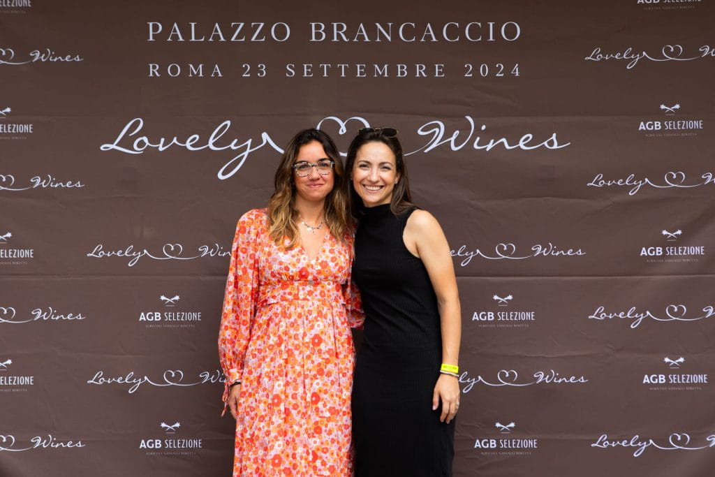 Due donne posano e sorridono davanti a uno sfondo con il marchio Lovely Wines durante un evento a Palazzo Brancaccio a Roma il 23 settembre 2024. Una indossa un abito floreale, l'altra un abito nero senza maniche.