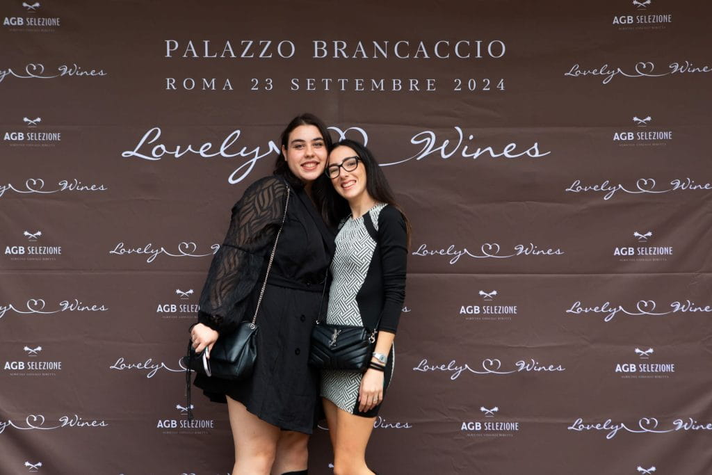 Due donne sono vicine e sorridono davanti a un fondale marrone con la scritta "Palazzo Brancaccio", la data dell'evento e "Lovely Wines". Entrambe indossano abiti e tengono in mano piccole borse.