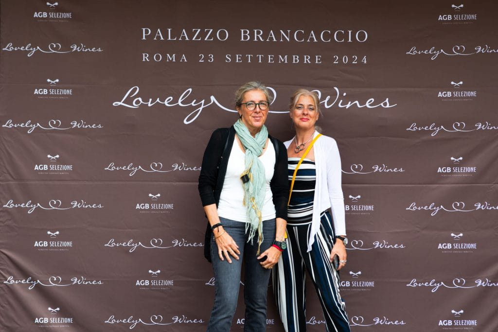 Due donne posano insieme davanti a un fondale brandizzato durante un evento a Palazzo Brancaccio, Roma, il 23 settembre 2024. Il fondale mostra gli sponsor dell'evento e il logo di Lovely Wines.