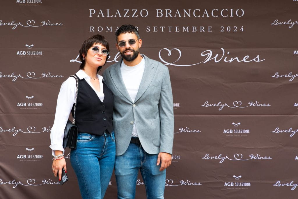 Due persone posano insieme, sorridenti, davanti a uno sfondo marrone con la scritta Palazzo Brancaccio Roma Settembre 2024 e Lovely Wines. Sono vestiti in modo casual e appaiono a un evento.