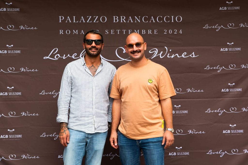 Due uomini sorridono davanti a un fondale marrone con testo bianco che promuove un evento enologico a Palazzo Brancaccio, Roma, nel settembre 2024. Uno indossa una camicia a righe, l'altro una maglietta gialla ed entrambi hanno gli occhiali da sole.